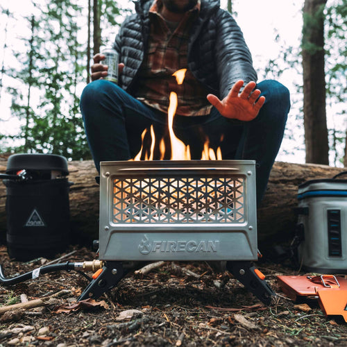 Ignik FireCan Elite portable propane fire pit with flames, person warming hands outdoors in forest
