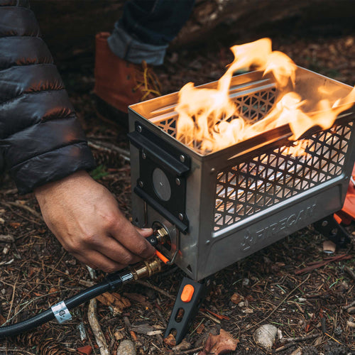 Ignik Outdoors FireCan Elite Fire Pit, a compact propane fire pit ablaze, user connecting hose in forest