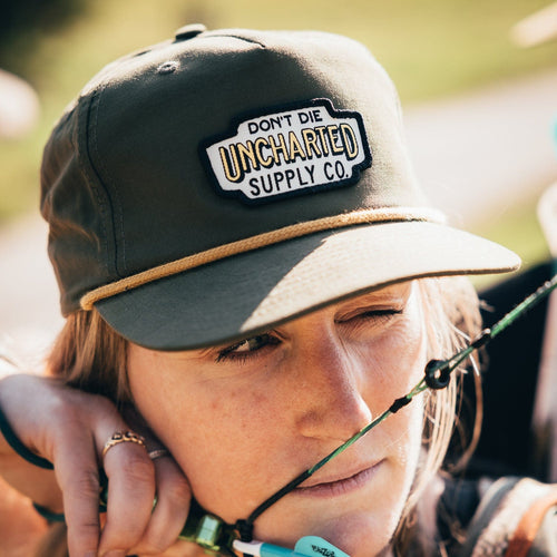 Green & gold Rover Hat worn by a woman aiming an arrow, featuring the Uncharted Supply Co patch