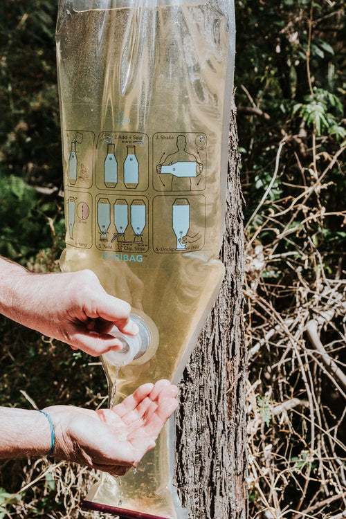 Puribag water purification system showing dirty water inside, dispensing clean water into a user's hand outdoors