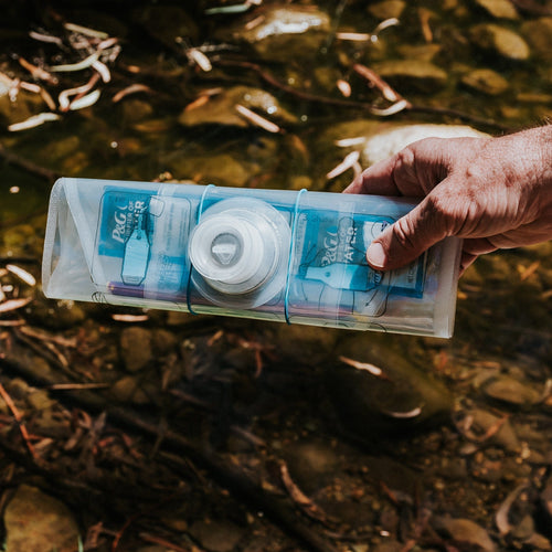 Puribag water purification system held by a man's hand over a stream, showing P&G label and filter cap for survival