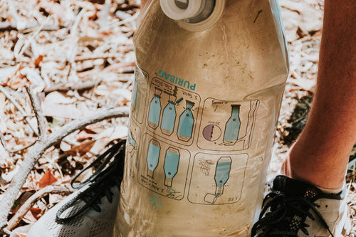 Puribag portable water purifier with instructions for use, held by a person in a dry outdoor setting