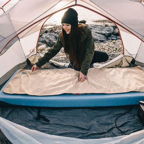Flipside Heated Bed Cover by Ignik Outdoors, woman setting up beige camping mattress warmer in tent