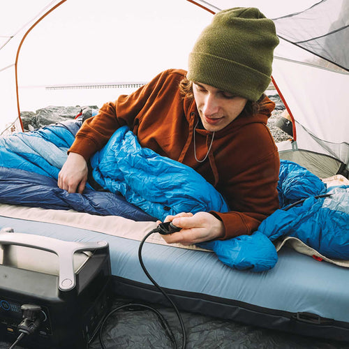 Flipside Heated Bed Cover by Ignik Outdoors heats a sleeping pad. A camper in a tent adjusts the portable mattress warmer.