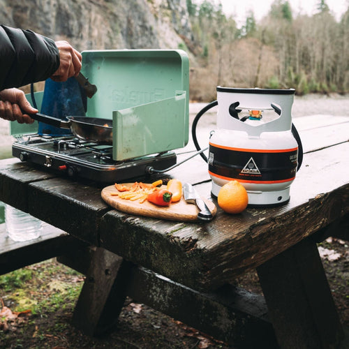 Ignik Gas Growler 3.8 Naked, a white portable propane tank, powers a camp stove for cooking outdoors on a picnic table