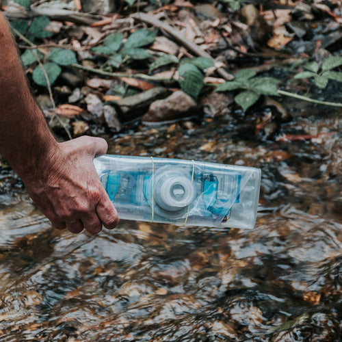 Puribag portable water filter held by a hand, collecting water from a natural stream in an outdoor setting