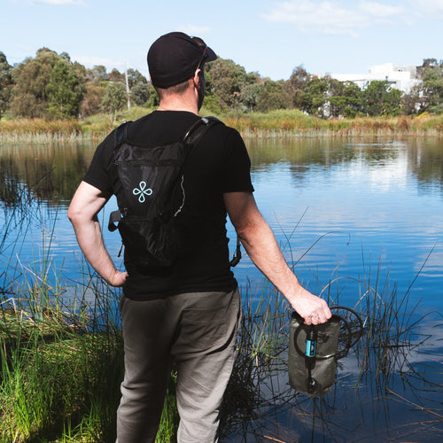 Puribag Adventure Pack: man wearing black hydration backpack holds olive green Puribag filter over lake