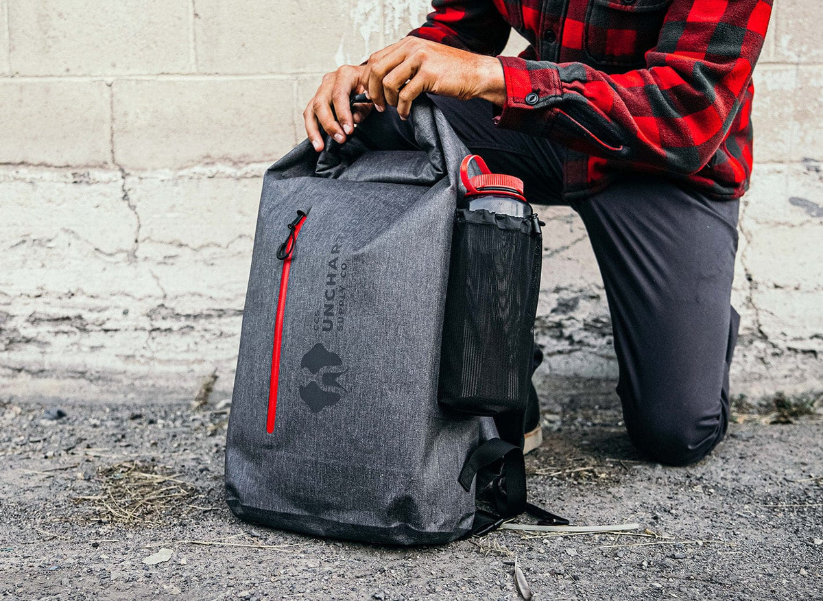 Person in plaid shirt securing a gray roll-top seventy2 dry bag, with a red zipper and water bottle