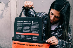 Woman examines a survival kit with integrated guide, showing steps for getting warm, starting fire, and first aid