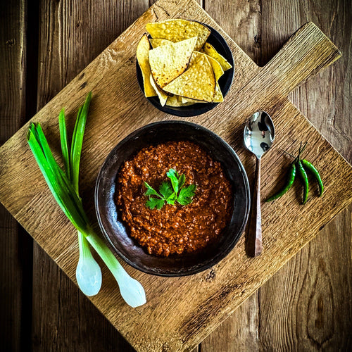 Essential Provisions BISON CHILI ready-to-eat meal in a dark bowl with tortilla chips, green onions, and chili peppers on a wood board