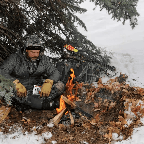 Person sitting by a fire in the snow with hunting gear and equipment around holding a possibles pouch