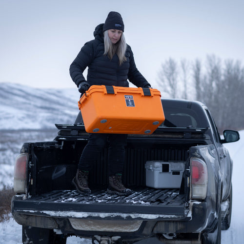 Person loading an orange basecamp 60L cooler onto a truck bed in a snowy landscape