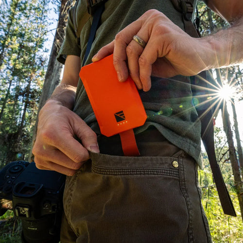 Dark Energy Poseidon Nano orange portable power bank slides into a hiker's pants pocket with a camera nearby and sun rays in a forest