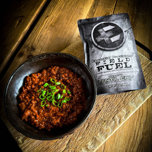 Lentil Chili ready-to-eat meal by Essential Provisions Field Fuel in a dark bowl, green onions, next to its pouch on wood