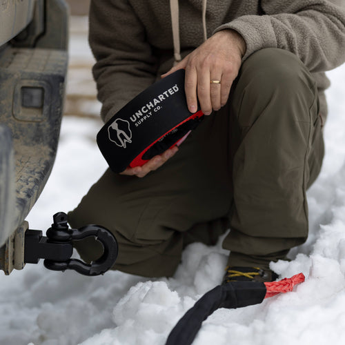 The Extractor - Tow Strap, black sleeve with red synthetic rope, held by person in snow by vehicle hitch & shackle