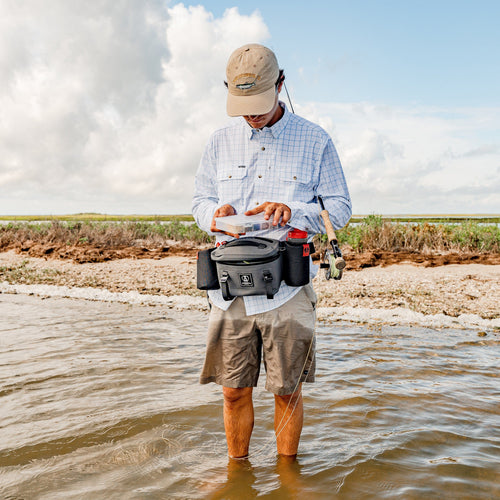Man standing in shallow water holding a fishing rod and wearing a fishing vest with tackle box and a park pack