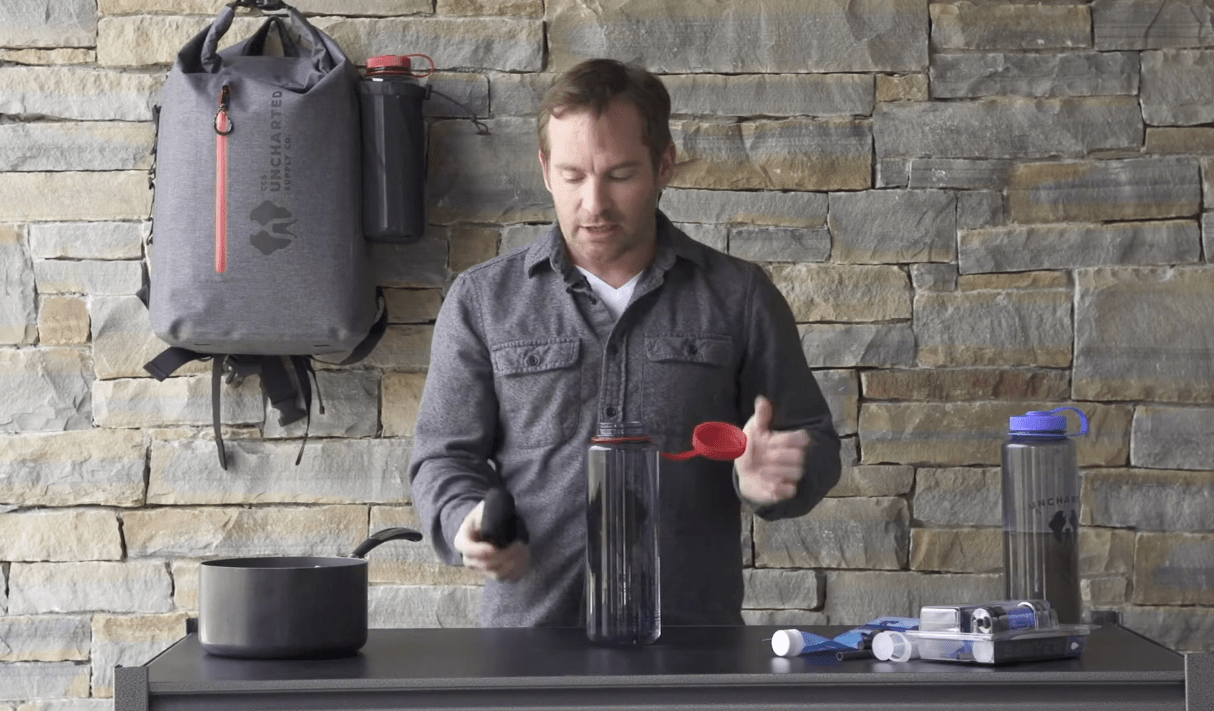 Uncharted Supply Co. water filter demonstration with man, clear bottle, and survival gear on table