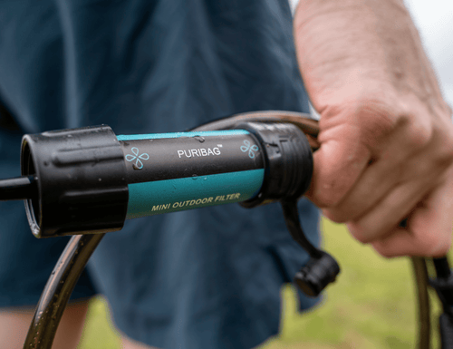 Puribag hydration bladder filter, turquoise and black, held by a hand with wet hoses outdoors