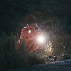 Tactical flashlight held by a woman in an emergency blanket, lighting a dark outdoor survival scene