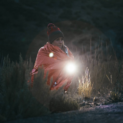 Tactical flashlight held by a woman in an emergency blanket, lighting a dark outdoor survival scene