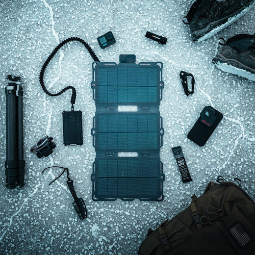 Dark Energy Spectre 18W portable solar charger unfolded on snow, charging a power bank, surrounded by rugged outdoor gear