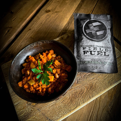 Essential Provisions VEGETABLE STEW MRE in a rustic bowl with herbs, beside its Field Fuel pouch
