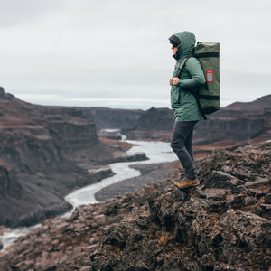 A person standing above a river canyon with an Olive 65L vault on their back