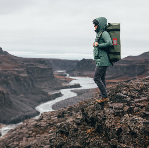 A person standing above a river canyon with an Olive 65L vault on their back