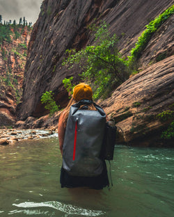 Hiker with Seventy2 Shell wading through a river in a majestic canyon with towering rock walls