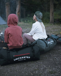 Two people sit on black and grey Uncharted vault duffel bags by a campfire, ideal camping gear for outdoor trips
