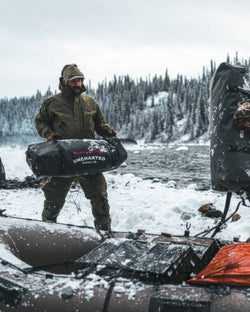 Man in winter gear carrying Uncharted Supply Co. vault 100L duffel bag by a snowy river