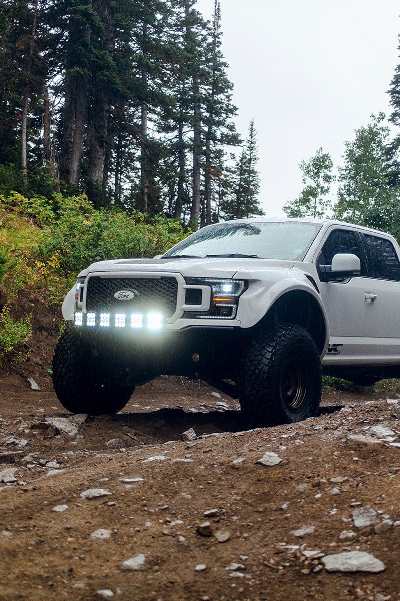 White Ford Raptor off-road truck with bright LED lights navigating a rocky forest trail