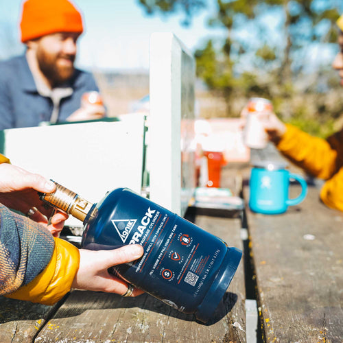 Ignik TapRack Refillable Propane System held in hands on a wooden table, people gathered outdoors