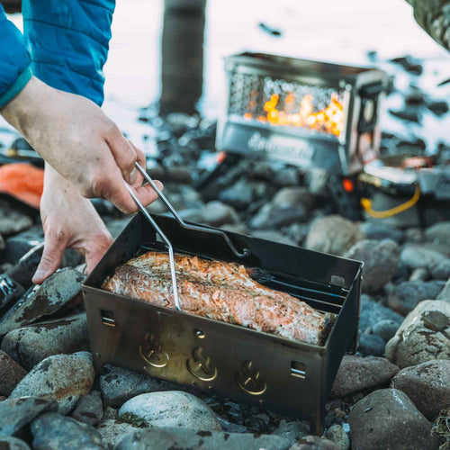 Ignik FireCan Deluxe Fire Pit grilling salmon outdoors on a rocky shore, hands adjusting food