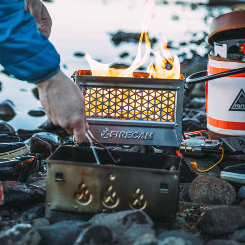 Ignik FireCan Deluxe Fire Pit glowing with flames, a portable propane fire pit on rocks. A hand tends the fire