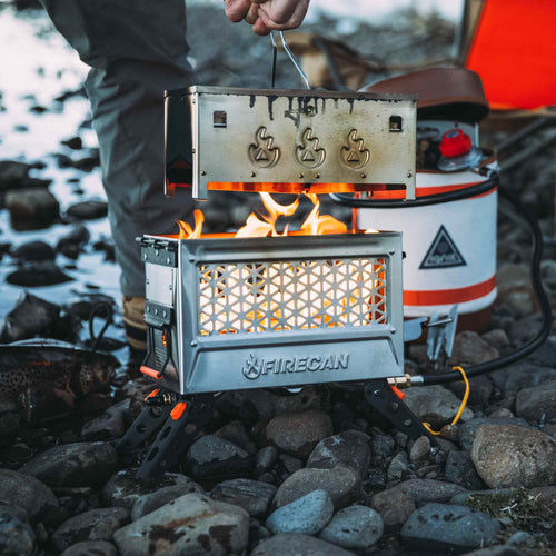 Ignik FireCan Deluxe Fire Pit, a portable fire pit with grill, showing flames on a rocky riverbank