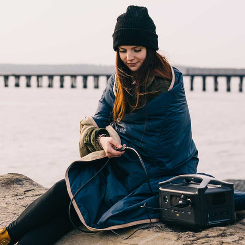 Topside Heated Blanket by Ignik Outdoors, woman wrapped in blanket plugging into a portable power station outdoors
