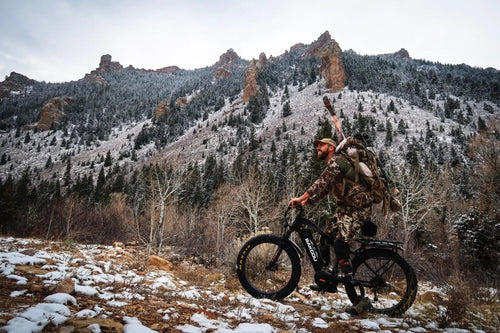 Bakcou Mule electric fat bike ridden by a hunter in camo, carrying gear and game through a snowy mountain landscape