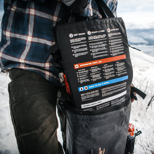 Person holding a black survival bag with instructions against a snowy background