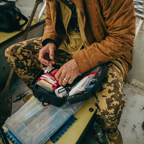 Person in camouflage pants and brown jacket organizing items in a first aid plus case on a boat.