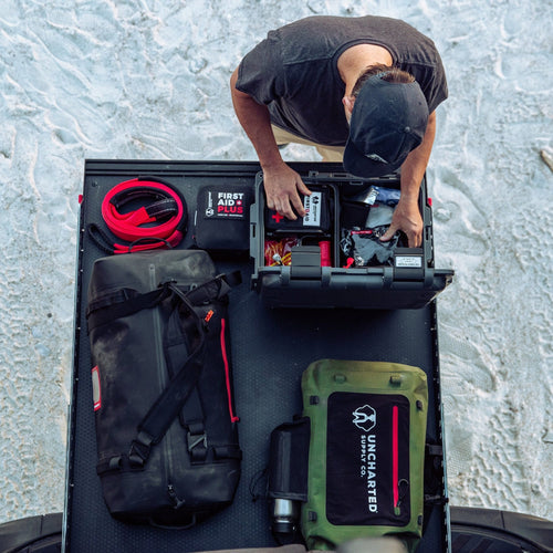 Person organizing a basecamp 30L survival kit next to other uncharted items