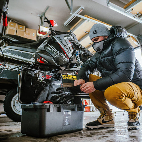 The Basecamp - 30L waterproof emergency kit, open with supplies, man putting on gloves beside snowmobile