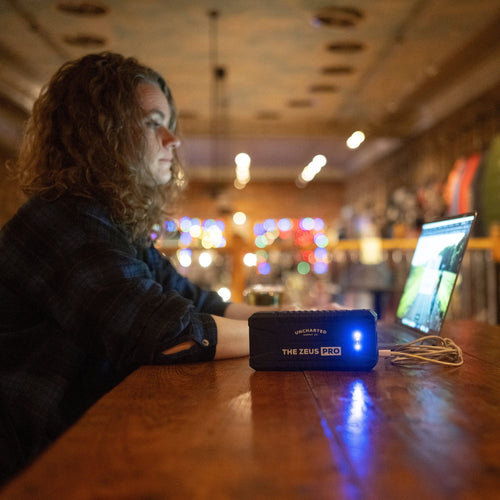 Person sitting at a bar with a laptop and 'The Zeus Pro' device on a wooden table.