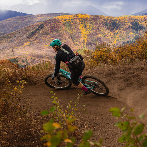 Person riding a mountain bike on a dirt trail with autumn-colored mountains in the background wearing a park pack