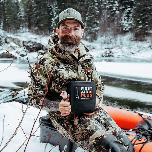 Man in camouflage clothing holding a First Aid Plus kit in a snowy outdoor setting.