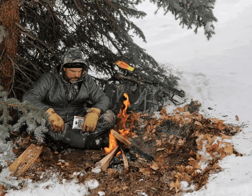 Person sitting by a fire in the snow with hunting gear and equipment around holding a possibles pouch