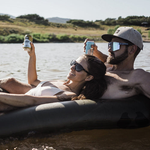 Two people relaxing in a rapid raft by a lake, holding drinks.