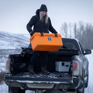 Person loading an orange basecamp 60L cooler onto a truck bed in a snowy landscape