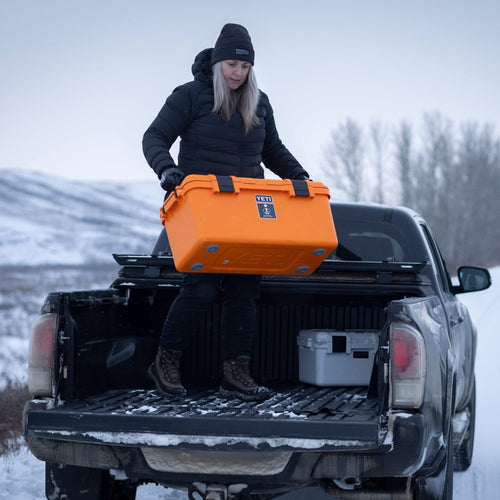Person loading an orange basecamp 60L cooler onto a truck bed in a snowy landscape