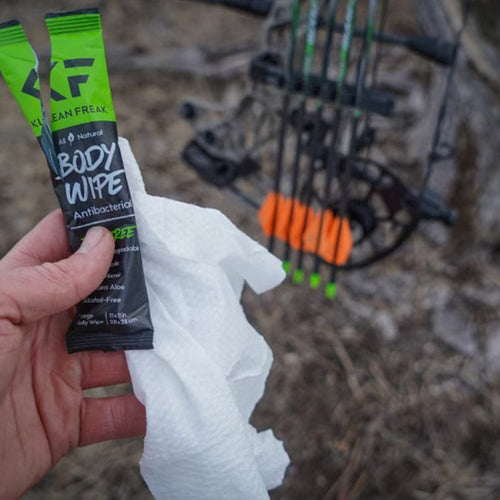 Klean Freak Body Wipes, two packets and an unfolded wipe held in a hand, with hunting bow & arrows in outdoor setting